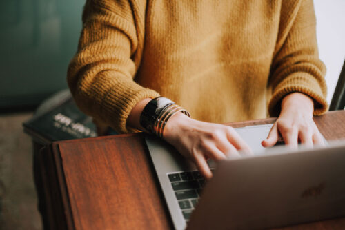 Eine Frau, die einen gelben Pulli trägt, arbeitet an einem Laptop. Man sieht nur ihren Oberkörper bis zu den Schultern.