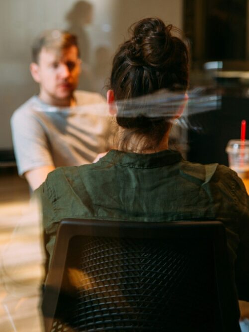 Zu sehen ist eine sitzende Frau mit Dutt von hinten an einem Tisch in eine Besprechungsraum. Ihr gegenüber sitzt ein unscharf zu erkennender Mann mit grauem T-Shrt.