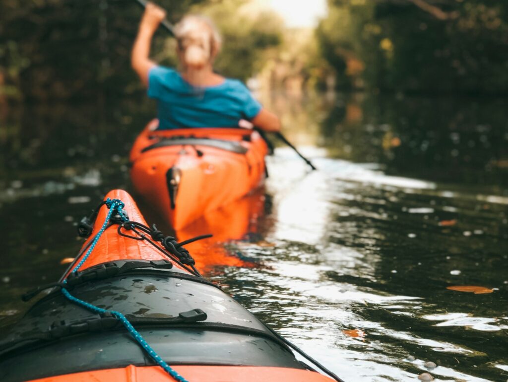 Die Spitze eines Kanus. Im Vordergrund unscharf zu erkennen eine blonde Frau, die im kanu paddelt.